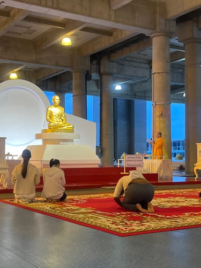 In Tha Khlong Town Municipality, Thailand, a Buddhist ceremony is underway. A golden statue of a seated monk, likely a revered figure, is prominently displayed on a white tiered platform. In front of the statue, three individuals are kneeling on a red carpet adorned with intricate patterns. Two women, facing away from the camera and towards the statue, are dressed in light-colored attire. A third individual, also kneeling and facing away, wears a white head covering and dark clothing. To the right, a monk in saffron robes stands at a table, holding an object and appearing to lead the ceremony. A sign with Thai script is placed on the table. The environment features large concrete pillars and a textured ceiling, illuminated by overhead lights, suggesting an indoor or covered outdoor space. The overall scene depicts a religious gathering with elements of reverence and tradition.