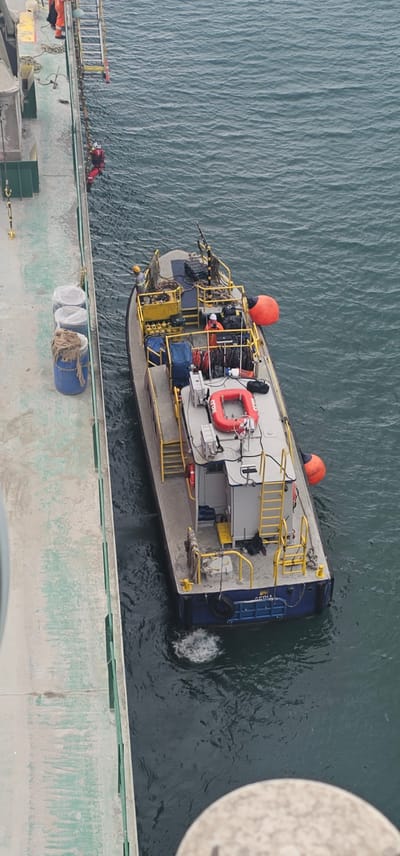 An aerial view shows a boat docked next to a pier in Cristóbal, Panama. The boat is small and appears to be a utility or work vessel, painted blue with yellow railings and ladders. There are several orange buoys attached to the side of the boat. On the deck of the boat, there are various pieces of equipment and supplies, including what appears to be a red life raft. Two individuals in orange safety suits are visible on the boat, one near the stern and another standing by a ladder. On the pier, there are three blue barrels, two covered with white plastic and one wrapped in burlap. Further down the pier, a person in a red life vest and red pants is descending a ladder into the water, presumably for a rescue operation. The water is dark and choppy, with white foam indicating the boat's propulsion. The pier itself is concrete with green painted edges and yellow safety markings. The overall scene suggests a maritime operation in progress.