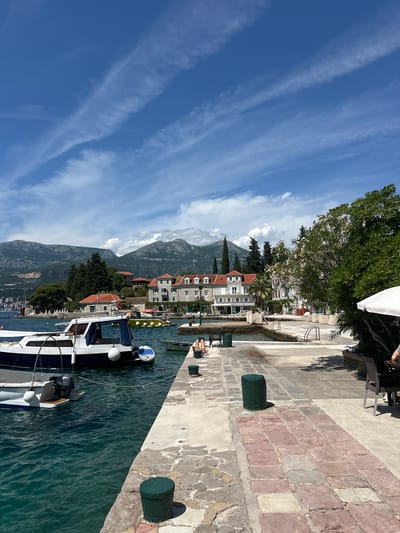 This image depicts a sunny coastal scene in Luštica, Montenegro, under a clear blue sky with sparse, wispy white clouds. In the background, large, rugged mountains covered in some vegetation rise prominently. Along the base of these mountains, particularly on the left, a settlement with numerous buildings is visible, extending up the hillside.
In the mid-ground, traditional stone buildings with red tiled roofs and multiple stories line the waterfront. Many feature green window shutters. Interspersed among these buildings and along the shore are various trees, including tall, slender conifers and broad-leafed trees, some bearing white or pink blossoms. On the far left, a section of beach with white lounge chairs and umbrellas is visible.
The foreground is dominated by a concrete pier or promenade with varied paving, including grey concrete, reddish square pavers, and irregular stone slabs. Dark green cylindrical bollards are spaced along the pier. The water is a clear, dark turquoise, exhibiting ripples from gentle movement.
Several boats are moored in the water. In the lower-left, a white and dark blue motorboat, marked "HN 145," is prominent, displaying white fenders and a Yamaha outboard motor. A smaller, light-colored boat is partially visible behind it. Further to the right, a yellow inflatable boat with a canopy is moored near the buildings. A small, light grey dinghy is also visible closer to the pier.
Two individuals are seated on the edge of the pier in the mid-right, with their legs dangling over the water; one is wearing light-colored clothing. On the far right, under a white umbrella, parts of a person's arm are visible at an outdoor seating area with wicker chairs and tables. A dark metal park bench is situated on the pier to the left, and metal handrails for water access are visible further along the pier on the right.
