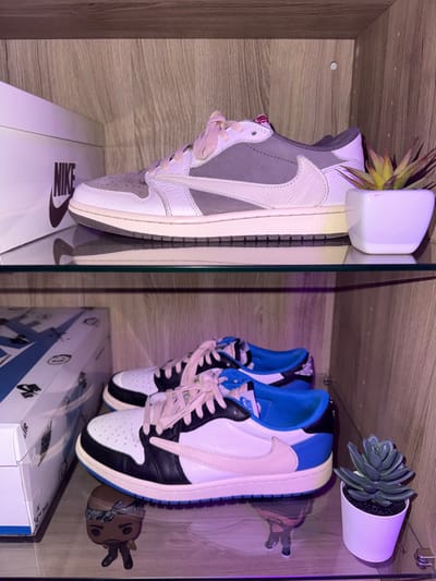 Inside a wooden shelf with glass doors, two pairs of Nike Travis Scott sneakers rest on their respective boxes. On the left, the "Neutral Grey" colorway is showcased alongside a small faux pineapple plant. On the right, the "Fragment" colorway stands with a small succulent and a toy figurine. The overall scene is illuminated with a slight purple hue, adding to the contemporary aesthetic.