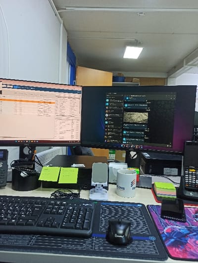 A cluttered desk features two monitors, a keyboard, a mouse on a patterned mousepad, and a mug labeled "CONS." Sticky notes are scattered around. A barcode scanner and other electronic devices are visible. The foreground is dominated by office equipment, while the background shows part of an office room. The scene gives an impression of a busy workspace.