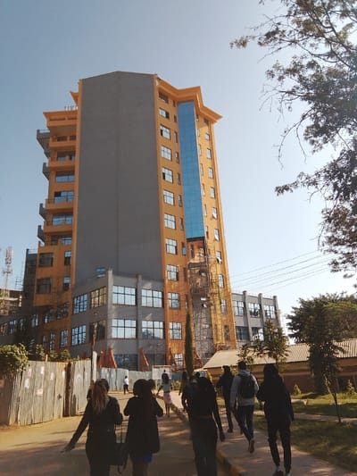 This image depicts a street scene in Karingani ward, Kenya, with a prominent multi-story building under a clear blue sky. The building features an orange-ochre facade, a large grey side panel, and a vertical section of blue reflective glass. Scaffolding is present on several exterior sections, indicating ongoing construction or maintenance. Approximately ten to fifteen individuals, mostly viewed from behind, are walking away from the camera on a paved pathway in the foreground. To the left, a corrugated metal fence borders the pathway, with a communication tower partially visible beyond it. On the right, mature trees with some bare branches are visible, and overhead, multiple electrical or communication lines traverse the frame. A small, single-story building with a brown roof is partially visible behind the trees on the right.