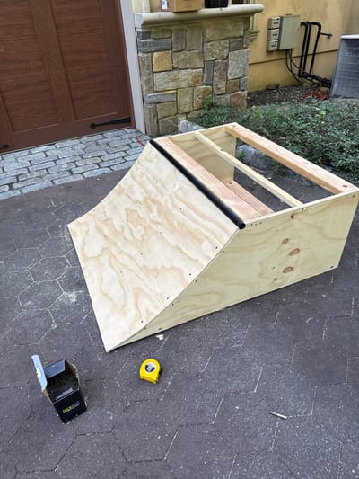 The image displays an outdoor scene on a paved area in Cresskill, United States, featuring a partially constructed wooden ramp. The ramp consists of light-colored plywood sheeting secured with screws, forming a sloped face and vertical sides, with exposed wooden structural framing visible at its top and rear. A black, cylindrical metal rail or pipe is affixed horizontally along the upper edge of the ramp's sloped face.

On the dark gray hexagonal paver ground in the foreground, an open black cardboard box containing numerous black screws, labeled "KREG FAST," is positioned to the left of the ramp. A yellow measuring tape is situated next to the screw box, and a single silver screw bit lies further right on the pavers.

The background features a building facade. To the left, a large brown wooden garage door with black decorative hinges is partially visible. To the right and behind the ramp, a section of a building wall includes a stone veneer column transitioning to a light-colored stucco surface. On this stucco wall, three electrical utility boxes and black conduit piping are mounted. A small, brown cardboard box rests on a ledge atop the stone column. Further to the right, an outdoor air conditioning unit with a metal grille is partially visible. Green ground cover plants are present in a landscaped bed between the paved area and the building wall. There are no individuals present in the scene.