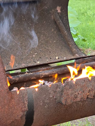 A close-up, waist-level shot shows a rusted metal barbecue grill with an active fire. The grill has a weathered, dark brown exterior with visible patches of rust and pitting. Flames are visible through a broken section of the grill's lower edge, indicating a fire within. Wisps of white smoke rise from the left side of the grill. In the background, blurred green grass and foliage suggest an outdoor setting, possibly a yard. The scene is devoid of any human presence. The location is noted as being in the city of Ермолаево, Russia.