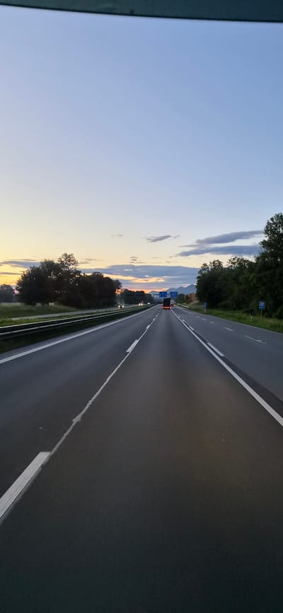 This vertical-format image captures a view from inside a vehicle traveling on a multi-lane highway near Raubling, Germany, during either dawn or dusk. The sky transitions from a light blue overhead to a band of warm yellow, orange, and faint pink hues at the horizon, with scattered grey clouds in the lower sky.
The asphalt highway features at least two lanes in the foreground, marked by white solid and dashed lines. A metal guardrail separates the lanes of travel from an opposing set of two lanes, which also feature a guardrail. In the same direction of travel, a red passenger car is visible in the left lane, and a dark-colored commercial truck is further ahead in the right lane, both displaying illuminated taillights. In the opposite direction, several vehicles are visible in the distance, indicated by their illuminated headlights.
Dense green trees and vegetation line both sides of the highway. On the right, several blue rectangular road signs are visible in the middle distance, along with a smaller blue sign closer to the foreground. Beyond the trees and signs on the right, distinct mountains or large hills are silhouetted against the colorful horizon. The very top edge of the image is obstructed by a dark, horizontal element, likely part of the vehicle's interior. No people are discernible.