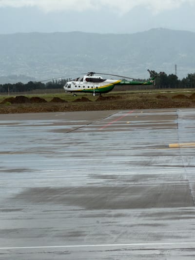 The image shows a helicopter parked on a wet tarmac in the city of Ianeti, Georgia. The helicopter is white, green, and yellow and marked "Border Police of Georgia" and the registration number "GBP-10005." The tarmac has visible lines and markings, including a red stripe. Beyond the tarmac is a grassy area with trees and a hazy mountain range in the background. The weather appears overcast.