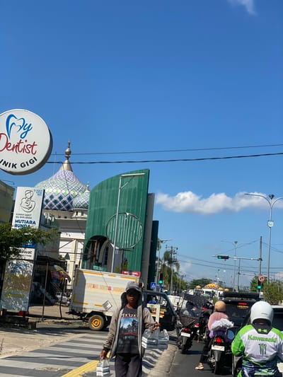 A street scene in Jawa, Indonesia, features a bright blue sky with scattered clouds. In the foreground, a person walks across a crosswalk, carrying two plastic bags of bottled water. They are wearing a gray hooded jacket over a graphic t-shirt and dark pants. Behind them, traffic moves along the road, including motorcycles, cars, and a white truck with "PT. ARTIKA SENTOSA INDONESIA" printed on its side. To the left, a sign for "My Dentist" and a billboard for "RUMAH SAKIT IBU DAN ANAK MUTIARA BUNDA" are visible. Further in the background, the distinctive domes of a mosque, adorned with green and white geometric patterns, punctuate the skyline. Streetlights and power lines crisscross the upper portion of the image.