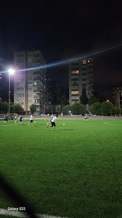 An outdoor, artificial turf soccer field is illuminated by stadium lights at night in the city of Çukurova, Turkey. The field is bordered by a fence and trees, with apartment buildings visible in the background. Several individuals, appearing to be young men, are engaged in a soccer game. One person in the foreground is running with the ball at their feet. Other players are scattered across the field, some running and others observing the game. The lighting casts a bright glow on the field, creating a lively atmosphere despite the darkness of the night sky. The "Galaxy S22" watermark is present in the lower left corner.