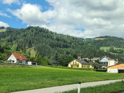 The image depicts a rural scene in the vicinity of St. Peter ob Judenburg, Austria. A gently sloping, well-maintained green lawn occupies the foreground. A paved road or path runs along the bottom edge of the frame. In the mid-ground, several houses are visible. To the left, a light grey house with a red-tiled roof and a detached garage is situated on a slight incline, bordered by a stone wall and well-kept hedges. Further to the right, a bright yellow house with multiple windows and a balcony stands, also surrounded by manicured greenery. Behind this, a collection of other residences with various rooflines and colors are nestled amongst trees. A large, densely forested hill or mountain forms the backdrop, stretching across the upper half of the image. The sky above is blue with scattered white, fluffy clouds, suggesting fair weather. The overall impression is one of a peaceful, residential area integrated into a natural, hilly landscape.