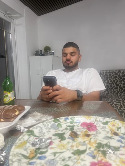 In a brightly lit room with white walls and a wood-paneled ceiling, a young man with short, dark hair and a beard is seated at a table, engrossed in his smartphone. He wears a white t-shirt and a digital watch on his left wrist. In front of him, a green glass bottle of lemon-flavored drink sits next to a plate of chocolate-covered pastries. A patterned couch is visible in the background, along with a white cabinet and a window. The table surface is dark and reflective, with scattered glitter. The scene appears to be a domestic setting, possibly a dining area or living room. The specific location is not discernible from the image alone, but if it is in Rosenovo, Bulgaria, it would be an indoor residence there.