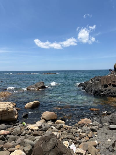 The image depicts a rocky coastline under a clear blue sky with scattered white clouds. The sea is a deep blue-green, with waves gently lapping against the shore. The foreground is dominated by a jumble of rocks of various sizes and colors, some smooth and rounded, others rough and angular. In the water, larger rocks are visible, some breaking the surface and creating small splashes. To the right, a black, columnar rock formation extends into the sea, with a white rope railing suggesting a pathway or viewing area. A sign is mounted on a post near the railing. The environment appears natural and somewhat rugged. There are no people visible in the image. The presence of some white and green litter on the rocks suggests human activity, despite the absence of people at the moment. The scene is a snapshot of a coastal landscape, likely a tourist spot given the railing and sign. The location is identified as Phú Lương, Vietnam.