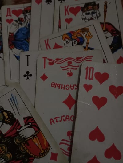 The image displays a close-up, top-down view of multiple scattered playing cards, consistent with a Russian "Atlas" pattern deck, featuring Cyrillic letters and distinct court card artwork. The cards are arranged in an overlapping, unorganized fashion on an indistinct, light-colored surface that serves as the background. Prominently visible cards include a Queen of Hearts, marked with the Cyrillic letter "Д" (Dama) and depicting a woman holding a red rose; a King of Hearts, marked with "К" (Korol) and illustrating a bearded man with a scepter; an Ace of Clubs, denoted by "Т" (Tuz) and a club symbol; and a 10 of Hearts, identified by the numeral "10" and multiple heart pips. Another partially obscured card shows a diamond suit and the inverted Cyrillic text "КАСНЫЕ АТЛАС" (Krasnye Atlas). The cards exhibit minor wear along their edges and surfaces, suggesting use. No individuals are present in the scene, nor are specific actions or interactions taking place other than the static arrangement of the cards. The specific location within Kurganinsk, Russia, is not identifiable from the provided visual information.
