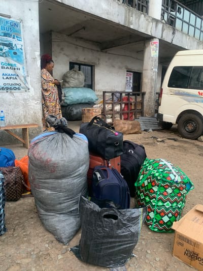 This is a medium shot, eye-level photograph taken outdoors at what appears to be a transportation hub in Elelenwo, Nigeria. The scene is characterized by a mix of luggage and people gathered near a building and a vehicle.

In the foreground, there is a large, grey, woven sack with red markings and a black knot on top. To its right, a large black plastic bag lies open on the ground. In front of the grey sack, partially visible, is a brown and black checkered duffel bag and an orange bag. To the right of the grey sack and slightly behind it is a large, black, zippered duffel bag. Further right and slightly behind is a dark blue suitcase. To the far right, a large, brightly colored green and red patterned bag is prominently displayed, secured with white straps. A cardboard box with printed text is visible to the bottom right.

In the mid-ground, a woman with red braided hair is standing, leaning against a concrete wall. She is wearing a long, patterned dress and is looking down at a white smartphone she is holding in her hand. Next to her, there are two large, light blue sacks, one stacked on top of the other. Behind the woman and the sacks, part of a building facade with a window is visible.

In the background, a white van is parked on the right side of the frame, its rear visible. Part of a corrugated metal fence or barrier is seen behind the van. A metal shelving unit filled with items is positioned against the building. The ground is rough and appears to be a dirt or gravel surface. A wooden bench is situated to the left of the woman. A blue plastic water bottle sits on the bench. A poster with images of vehicles and text is affixed to the wall on the left. The overall environment suggests a busy, utilitarian area, likely a bus station or cargo loading point.