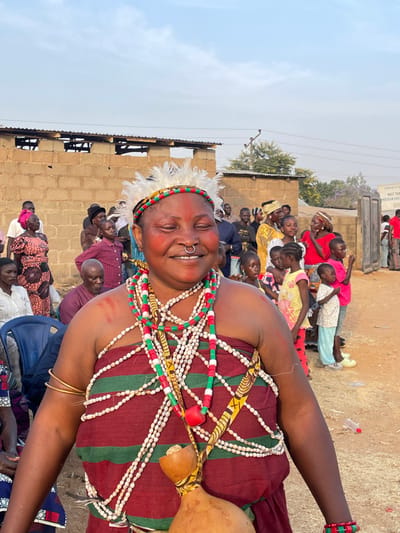 The image captures a vibrant outdoor gathering, with a focus on a smiling woman adorned in traditional attire. The photograph is oriented vertically, showing the woman prominently in the foreground.
The central figure is a woman with dark skin, broad features, and a joyful, wide smile. Her eyes are closed in her expression of happiness. She has red markings on her cheeks and a metal septum piercing in her nose. Her head is decorated with a white feathered headpiece secured by a band of red, green, and yellow patterns. She is heavily adorned with multiple strands of beads; some are small white beads, while others are larger red and green cylindrical beads, creating a rich, layered effect across her chest and shoulders. Her attire includes a fabric garment, possibly a wrapped cloth, featuring alternating wide stripes of dark red/maroon and thinner stripes of green with yellow patterns. A light brown gourd is visible on her right side, attached to her clothing. She wears simple gold-colored bangles on her left wrist and bracelets made of red and green beads on her right wrist.
Behind the woman, a crowd of people, including many children and adults, are gathered. They appear to be seated or standing, observing an event or performance. Several children are visible in the mid-ground, some looking in the same direction as the main subject, others glancing towards the camera. The setting appears to be a rural or semi-urban area, with simple buildings constructed from concrete blocks forming the backdrop. The ground is bare earth or dirt. In the upper right background, a faded sign is partially visible, on which the words "AN CHU" can be deciphered, likely part of "CHURCH". A tree with purple blossoms can be seen in the distance, and utility poles and wires crisscross against a light, clear sky, indicating daytime. The overall atmosphere conveyed by the main subject's expression is one of festivity and joy.