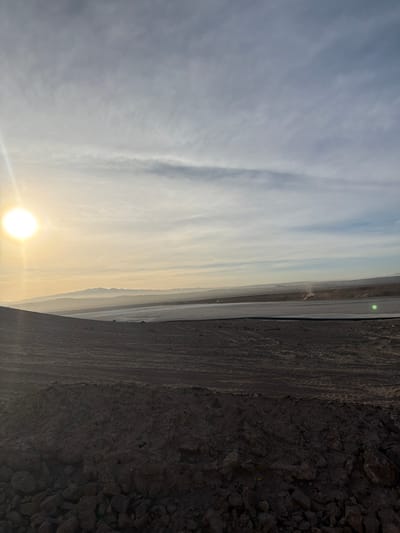 The image displays an arid, open landscape in Sierra Gorda, Chile, under a sky suggesting early morning or late afternoon light. The foreground consists of dark brown, uneven terrain composed of rocks and disturbed earth, presenting an irregular, excavated appearance. Beyond this foreground, a vast, flat, light-toned surface stretches horizontally across the midground, resembling a large industrial pad or a tailings pond. In the far distance, a faint, low mountain range is discernible through atmospheric haze. The sun is visible as a bright, luminous sphere in the lower left quadrant, casting a golden-yellow light across the left side of the scene and into the sky. The sky transitions from a bright yellow-orange near the sun to a lighter blue on the right, with scattered, wispy clouds throughout. A distinct green lens flare is present in the mid-right portion of the image, superimposed over the flat surface. A small, light-colored plume of dust or smoke rises from the flat ground in the mid-right. No human figures or discernible vehicles are present.
