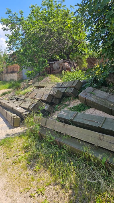 The image depicts an outdoor scene in Slatina, Bulgaria, with a large pile of weathered, olive-drab wooden crates stacked haphazardly on a gravelly slope. The crates appear to be old ammunition boxes, some showing signs of wear and tear, with metal hinges and latches. They are arranged in multiple layers, some overlapping, suggesting they have been stored or discarded.
The environment is somewhat overgrown with tall green grass and weeds growing around and between the crates. In the background, a dense green tree with a thick trunk dominates the upper portion of the image. To the left of the tree, a brick building with a grey metal door is visible, partially obscured by foliage. A chain-link fence runs along the property line in the background. The sky above is blue with scattered white clouds. The lighting suggests it is a bright, sunny day. There are no people visible in the image.