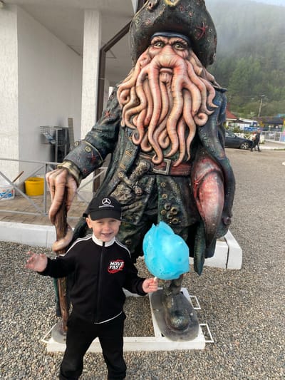 A young boy wearing a black tracksuit and a black baseball cap smiles while holding a blue cotton candy. He stands in front of a large statue of Davy Jones, a fictional character from Pirates of the Caribbean, which appears to be made of weathered materials. The statue is detailed, featuring tentacles on its face and a pirate hat. The background shows a building with white walls, a gravel ground, and a misty forest-covered mountain range. The scene appears to be outdoors, possibly in a tourist area.
