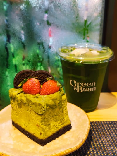 A slice of matcha cheesecake is served on a light-colored, speckled plate with a textured brown rim, sitting on a woven placemat. The cheesecake is square-shaped with a dark brown crust and a vibrant green filling topped with swirls of green frosting, two whole strawberries, and two halves of an Oreo cookie. To the right of the cheesecake is a clear plastic cup filled with a green beverage, likely iced matcha, bearing the white logo "Green & Bean". The background is a blurred green, suggesting a backdrop of plants or foliage, with condensation visible on a surface, possibly a window or partition. The scene appears to be indoors, perhaps at a cafe in Sakhu, Thailand, given the context.