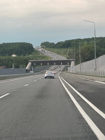 The image captures a multi-lane highway in Кощаково, Russia, on an overcast day. The perspective is from the driver's seat of a vehicle traveling on the road. In the foreground, the asphalt road is marked with white lane lines, indicating a clear path forward. To the right, a tall, sound-absorbing barrier runs alongside the highway, composed of grey panels. Further to the right, a streetlamp stands prominently.
The highway curves and ascends into the distance, flanked by lush green trees and rolling hills on both sides. A modern pedestrian bridge, featuring a dark, patterned surface, spans the highway in the mid-ground. Several cars are visible on the highway, some ahead and some in the opposite direction, navigating the road. One white car is particularly noticeable in the lane directly ahead. In the background, more cars are seen on the higher sections of the road, suggesting a flow of traffic. The sky is predominantly grey and cloudy, casting a muted light over the entire scene. Distant houses are nestled amongst the trees on the left side of the highway.