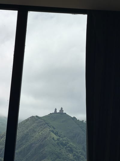 The image shows a view through a window frame of Gergeti Trinity Church, located in Stepantsminda, Georgia. The church is situated on top of a lush green mountain under a cloudy sky. The mountain has a steep, rocky incline covered in vegetation and scattered trees. The church itself appears to be a stone structure with multiple domes. The foreground is dominated by the dark frame of the window, with a dark curtain visible on the right side. The overall impression is a dramatic and picturesque landscape.