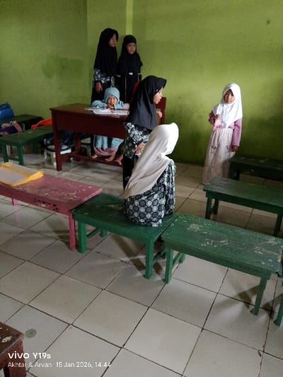 Inside a classroom with green walls and a tiled floor, several young girls are visible. Most are wearing hijabs, with one in a light blue hijab seated at a desk. Two girls in black hijabs stand behind the desk, while a girl in a white hijab stands to the side. Another girl, also in a white hijab, is kneeling on a green bench in the foreground. Other green and pink benches are scattered around the room.