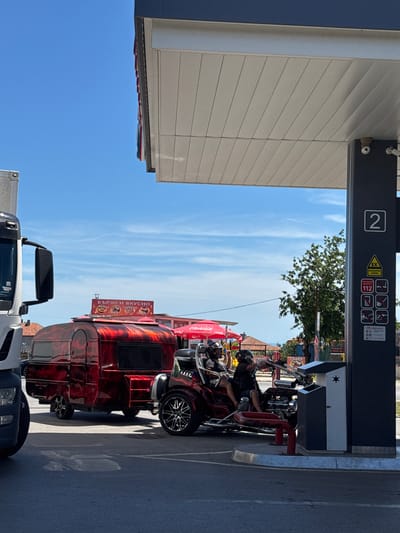 At a gas station in Byala, Bulgaria, under a clear blue sky, a red, flame-patterned trailer is parked. Next to it is a three-wheeled vehicle with two people on it, both wearing helmets. The gas pump, labeled with the number "2", is visible in the foreground. The trailer has a sign on top that reads "БЪРЗО И ВКУСНО", which translates to "Fast and Delicious". A large truck is parked on the left side of the image. The surrounding area appears to be a mix of commercial establishments and residential buildings.