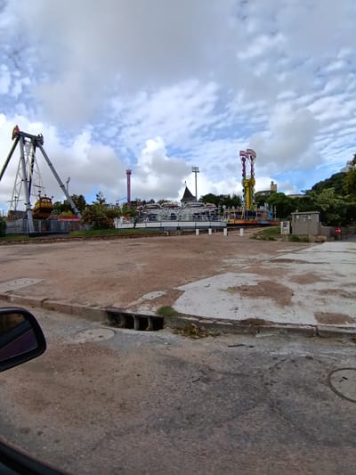 The image depicts a closed amusement park in Montevideo, Uruguay. Visible rides include a pirate ship, a drop tower, and a spinning ride. The park appears deserted, with no people visible. The ground is a mix of dirt, concrete, and paved road. A car side mirror is visible in the lower left corner of the frame. The sky is cloudy. A storm drain is visible along the curb in the foreground.