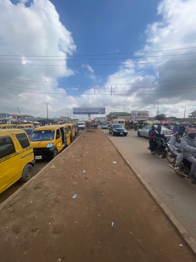 The image depicts a busy street scene in Ojodu, Nigeria, characterized by heavy traffic congestion. Numerous yellow minibuses ("danfos") are lined up, indicating a public transportation hub or waiting area. A mix of other vehicles, including cars and motorcycles, navigate the road, contributing to the congestion. Many people are on motorcycles. Several overhead power lines are present against a cloudy sky. A large sign for "SPRINGFORTH SCHOOL" is visible, suggesting the proximity of a school. The foreground shows a dirt/paved area.
