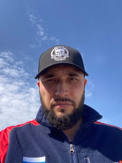 A man wearing a black cap with a white logo and a dark blue jacket with red sleeves is visible. The background is a bright blue sky with some scattered white clouds. The man has a beard and is looking directly at the camera. The perspective is from a low angle, looking up at the man. There is no information to confirm the location in Luzhitsy, Russia.