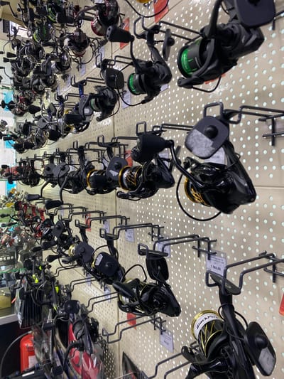 The image shows a wall-mounted display of fishing reels in a retail store. The reels are of various sizes and colors, with many featuring black and metallic finishes. Some reels have green lines on their spools. Several price tags are visible, with prices like 1,566.00 and 692.00. The display is set against a beige pegboard background with numerous circular holes. The overall scene depicts a commercial environment focused on sporting goods. The location is identified as Ivano-Frankivsk, Ukraine.