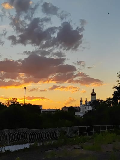 The image depicts a sunset over Korosten, Ukraine. In the foreground, a decorative metal fence runs horizontally across the frame, with lush green foliage visible behind it. A dark, uneven ground surface occupies the very bottom of the image. In the middle ground, a line of dark trees partially obscures a street with some buildings. Dominating the background are silhouetted trees and a prominent white church with golden domes and crosses. The sky is a gradient of pale blue at the top, transitioning to vibrant oranges and yellows near the horizon, illuminated by dramatic, colorful clouds. A single, dark bird is visible in flight against the sky. A cellular tower stands tall amidst the trees to the left of the church.