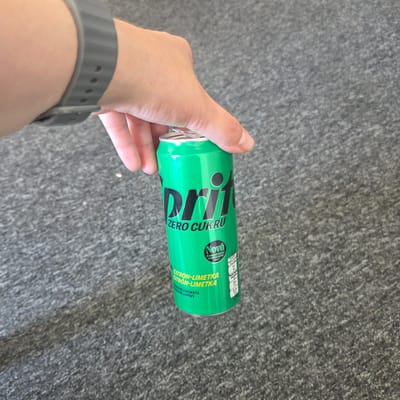 A hand wearing a grey fitness tracker is holding a green can of Sprite Zero Cukru. The can has black lettering for the Sprite logo and smaller text that reads "ZERO CUKRU". Below that, there is a small circle with "NOVÁ" in white lettering. Further down, the can indicates the flavor as "CITRON-LIMETKA" and "CITRÓN-LIMETKA", with the text "PŘÍRODNÍ AROMATA" and "PRÍRODNÉ AROMY" below that. The can is positioned over a grey, textured surface, likely a carpet. The image appears to have been taken indoors.