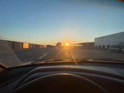 The image is taken from the interior of a vehicle, looking out through the windshield at a sunrise. The sun is low on the horizon, casting a bright, golden light across the scene. In the distance, a road stretches ahead, flanked on the left by a concrete wall topped with barbed wire, and on the right by a large industrial building. Trucks are visible parked near the building. The sky is a clear, pale blue, with a few small specks scattered across it, possibly dust or lens flare. The foreground shows the dashboard and steering wheel of the car, indicating the perspective of someone driving. The overall impression is one of a beginning to the day, possibly in an industrial or commercial area on the outskirts of Sviatopetrivske, Ukraine, given the architecture and the open, somewhat bleak landscape.