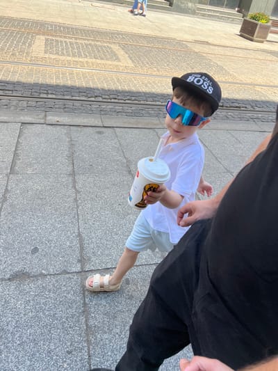 A young boy wearing a white polo shirt, light blue shorts, beige sandals, a black baseball cap that reads "THE REAL BOSS," and reflective sunglasses walks on a cobblestone sidewalk next to tram tracks. He is holding a white paper cup with a straw and a gold emblem, possibly from a fast-food establishment. A person's legs and torso, clad in black pants, are visible in the foreground on the right side of the image. The scene appears to be set in an urban environment, with a planter box containing greenery visible in the background, consistent with a location like Katowice, Poland. The lighting suggests it is daytime.