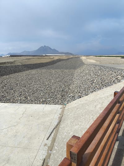 The image depicts an engineered waterway or canal lined with riprap in Huanchaco, Peru. The view is from a concrete structure, possibly a bridge or elevated walkway, with a partially visible wooden railing in the foreground. The canal bed is densely covered with light-colored stones that transition to darker stones further away. Beyond the canal, the landscape is predominantly flat and arid, marked by rows of low-lying structures that appear to be agricultural or industrial facilities. A large mountain with multiple peaks is present in the background, under a sky of scattered clouds. The overall scene conveys an environment managed for flood control or water diversion in a semi-desert region.