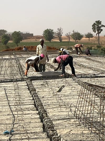 The image captures an outdoor construction scene on a bright, likely sunny day. Several individuals, presumably construction workers, are engaged in laying reinforcing steel bars (rebar) on what appears to be a large concrete slab or foundation under construction.
In the foreground and midground, approximately five to six men are visible, most of whom are bent over or stooping, actively working with the rebar. One prominent figure in the center-left wears a light-colored, possibly beige, long-sleeved shirt or tunic, dark trousers, and a light-colored cap. To his immediate right, another worker in a pink shirt and dark trousers is leaning forward over the rebar, appearing focused on the task. Other workers are similarly positioned, handling or adjusting the intricate grid of metal bars.
The construction surface is heavily covered with a dense network of rebar, arranged in rows with smaller, U-shaped or rectangular ties forming a continuous pattern. Larger, pre-fabricated rebar cages, forming square or rectangular shapes, are also visible, particularly in the upper right portion of the image. A light-colored, flat tool, possibly a trowel or concrete float, rests on the rebar grid in the central part of the image.
The background reveals a semi-arid or savanna-like landscape under a bright, hazy sky. Scattered trees, including some tall palm-like trees on the left, and smaller green bushes are visible. The ground in the distance appears to be dry, uncultivated earth or fields. The overall setting suggests a construction project in an open, possibly rural or developing area, under clear and bright daytime conditions.