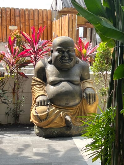 A statue of a seated Buddha is positioned outdoors in Singakerta, Indonesia. The statue is made of dark gray and gold-colored stone. The Buddha figure is depicted with a smiling face, a large belly, and prayer beads in his hand. It is surrounded by red and green tropical foliage, including some large broad-leaved plants at the top and red-leaved plants on the left and bottom of the frame. A wall made of bamboo sections is visible behind the Buddha. The ground where the statue is placed is tiled.
