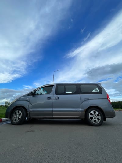 A gray Hyundai Starex minivan is parked on the side of an asphalt road in Бавыкино, Russia. A lamppost stands next to the vehicle. The sky behind the minivan is partly cloudy with blue patches visible. The area behind the van appears to be a grassy field with some trees in the background. A red and white striped curb is visible along the edge of the road.