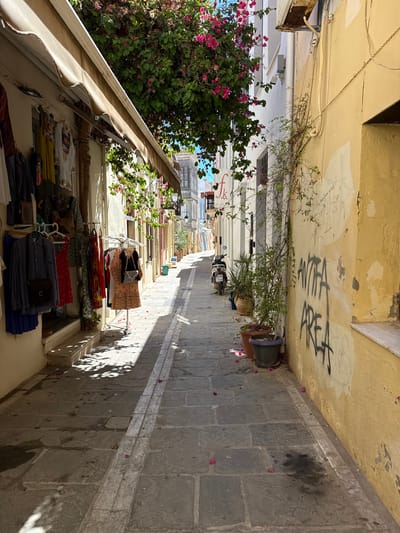 The image depicts a narrow, sun-drenched alleyway in the old town of Rethymno, Greece. The street is paved with large, irregularly shaped stone slabs, with a distinct, lighter-colored stone border running down the center. On the left side of the alley, a shop displays clothing on racks and hangers, with a beige awning extending overhead. The clothing includes dresses, shirts, and skirts in various colors and patterns. A small step leads up to the shop entrance.
The right side of the alley is dominated by a textured, yellow-painted wall adorned with graffiti. The graffiti, written in black spray paint, appears to read "AVTIPA AREA". Green vines climb the wall, and several potted plants, including succulents and other greenery, are placed along the base of the wall.
In the background, the alley recedes into the distance, revealing more buildings with architectural details characteristic of Greek old towns, including balconies and shuttered windows. A white scooter is parked on the right side of the alley, near the potted plants. Pink bougainvillea flowers cascade from unseen trees overhead, adding splashes of color to the scene and casting dappled shadows on the pavement. Scattered pink petals are visible on the ground throughout the alley. The overall impression is one of a vibrant, historic urban environment on a bright, sunny day.