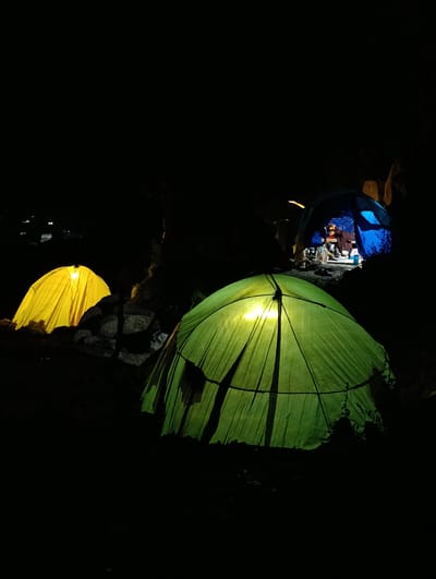In the city of Palchan, India, a nighttime scene unfolds with three illuminated tents set against a dark, natural environment. In the foreground, a large green tent is brightly lit from within, its fabric taut and revealing the tent's internal structure. To the left, a yellow tent is also illuminated, casting a warm glow. In the background, a blue tent is visible with people inside, gathered around a light source. Bottles and other items are scattered within the blue tent. The ground appears uneven and rocky, with some vegetation. Distant lights suggest the presence of a settlement.