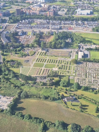 This is an aerial, high-angle photograph of a suburban area with residential housing, a cemetery, and a sports field. The image appears to be taken during daylight hours. In the foreground, a field with tall grass is visible, bordered by trees. A small, modern-looking house with a dark roof and light-colored walls is situated on a well-maintained lawn, surrounded by trees. A yellow line demarcates the boundary of the property.
Further into the image, a large cemetery is a prominent feature, with rows upon rows of headstones. Adjacent to the cemetery are several paved areas with parking spaces, and a large building, possibly a community center or facility for the cemetery. Beyond this, a football (soccer) field with white markings is clearly visible, alongside more residential buildings and wooded areas.
In the background, a densely populated urban area with various building heights and architectural styles can be seen, interspersed with green spaces and trees. Construction activity, indicated by earthmoving equipment and piles of materials, is evident in one section of the residential development. The overall scene depicts a blend of urban development, recreational spaces, and burial grounds, characteristic of a mixed-use suburban landscape. The presence of distinct housing developments, a large cemetery, and a sports field suggests this is likely a residential area on the outskirts of a city. Based on visual cues such as architectural styles and the planning of the residential areas, this location is consistent with the city of Dublin, Ireland, potentially an area like Glasnevin or a similar suburb with extensive green spaces and a large cemetery.
