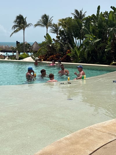 In this image, several people are enjoying themselves in a swimming pool at a resort in Punta Sam, Mexico. The water appears clear and a light turquoise color, with sunlight creating rippling patterns on the pool floor. The pool itself has a light-colored concrete edge.

In the foreground, a man wearing a blue baseball cap is standing in the water, holding a yellow drink. Further back, other individuals are visible, some with drinks as well. One person in the middle of the pool is partially submerged, and another is standing deeper in the water. The background features lush tropical vegetation, including palm trees and large-leafed plants, suggesting a warm, sunny climate. In the distance, beyond the pool and foliage, a glimpse of the ocean and thatched-roof structures can be seen, characteristic of a beach resort.

The overall atmosphere seems relaxed and festive, with people enjoying a casual outing at a tropical vacation spot. The lighting suggests it is daytime, likely late morning or early afternoon, with bright sunshine. There is no visible text in the image. The image is oriented vertically, with the top of the scene appearing on the left side.