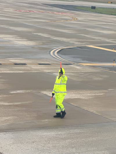 Aviation traffic controller at San Antonio International Airport in San Antonio, Texas, signals an aircraft on the tarmac. The controller wears a neon yellow safety suit, black gloves, and boots. They hold orange wands in both hands. The tarmac shows yellow lines, markings, and a water-like surface.
