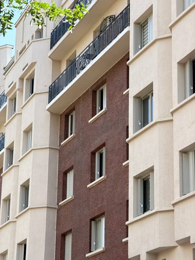 An exterior, eye-level, medium shot captures the side of a modern, multi-story apartment building in Courbevoie, France. The building features a staggered facade with alternating sections of light-colored, textured stucco and dark red brick. Black wrought-iron balconies with decorative patterns punctuate the upper floors. Numerous windows, most with reflective glass, are visible across both material sections, some appearing slightly ajar. Green foliage from a tree drapes down from the top left corner of the frame. The architecture suggests a residential area, likely in an urban setting. The sky is a light blue.