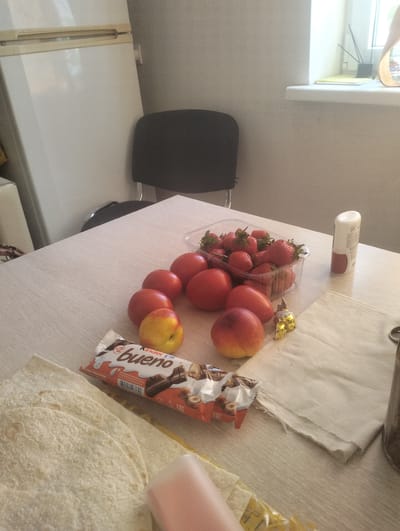 The image depicts a table with several food items, including strawberries in a plastic container, tomatoes, a peach, a Kinder Bueno chocolate bar, and what appears to be flatbread or lavash. A roll-on deodorant and a golden-wrapped candy are also present. In the background, a white refrigerator and a black chair are visible, along with a windowsill with a window and some decorative items. The scene suggests an indoor setting, likely a kitchen or dining area, possibly in a residential environment. The lighting indicates it is daytime. Without further context or identifying markers, it is impossible to ascertain the specific location in Smolensk, Russia.