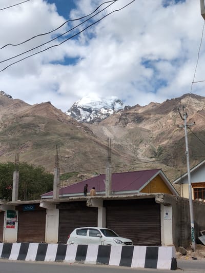 The image shows a street scene in Padum, India, with a snow-capped mountain range in the background. In the foreground, there are several shops with rolled-down shutters, a white car parked on the street, and a concrete barrier. Two men are standing on the roof of one of the shops, observing the surroundings. Power lines are strung across the sky, and a utility pole is visible on the right side of the image. The buildings appear to be constructed with concrete and metal roofing. The mountain slopes are rocky and barren, with patches of green vegetation. The sky is partly cloudy, with patches of blue visible. The overall impression is a blend of urban infrastructure and natural landscape.