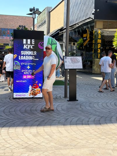 In Ashdod, Israel, at the Babylon Park, a man wearing a white t-shirt, white shorts, and a beige baseball cap stands near a large LED screen. The screen displays "KFC SUMMER LOADING..." along with an image of a KFC meal. To the left of the man, a young person in a white t-shirt and black shorts walks away from the screen, carrying a white shopping bag. In the background, other individuals are visible, and the architecture of the Babylon Park, including a building with dark, angular facade, is apparent. The ground is paved with interlocking stone tiles.