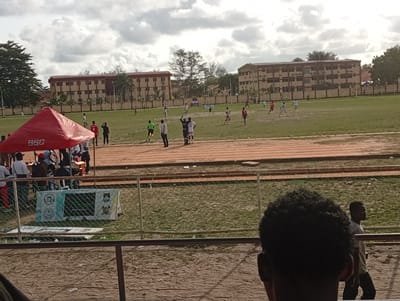 The image depicts a soccer match underway on a green field, viewed from what appears to be stadium seating. In the foreground, a fence separates the seating area from the field. Two individuals are partially visible in the foreground; one with dark, curly hair occupies a significant portion of the lower right corner. Behind the fence, a running track surrounds the soccer field. A red canopy tent is set up on the side of the field near the track. Several people are gathered around the tent, and a sign is placed in front of it. In the background, two multi-story buildings with repeating window patterns flank the field. The sky is overcast with heavy clouds.
The location is Ijanikin, Nigeria.