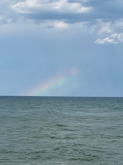 A distant, faint rainbow arcs across a pale blue sky with scattered gray clouds. Below the sky, a dark, choppy ocean stretches to the horizon. The water is a deep greenish-gray, with whitecaps indicating moderate waves. The scene appears to be from the coast of Sanzhiika, Ukraine, given the typical weather patterns and coastal landscape. There are no people or man-made objects visible in the frame.