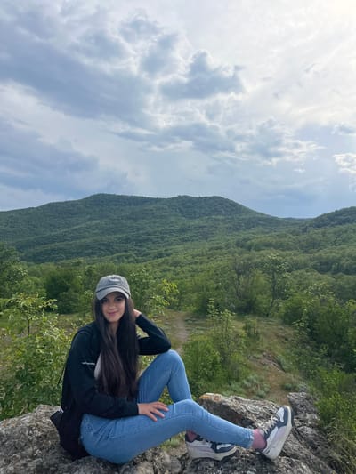 A young woman is seated on a rocky outcrop, seemingly in a mountainous or elevated terrain. She is wearing light blue jeans, a black jacket over a white top, and grey and white sneakers with pink socks. She has a grey baseball cap on her head and long, dark hair. The background reveals a lush, green mountain range, with a cloudy sky overhead. The location may be in the vicinity of Kamchia, Bulgaria, given the landscape details. The woman is looking at the camera and smiling gently, with her legs crossed. She is posing against the scenery, suggesting a leisurely activity or photoshoot amidst a natural setting.